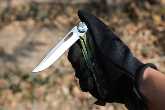 A gloved hand holding an open multitool with a green handle, blade deployed, on a forest floor